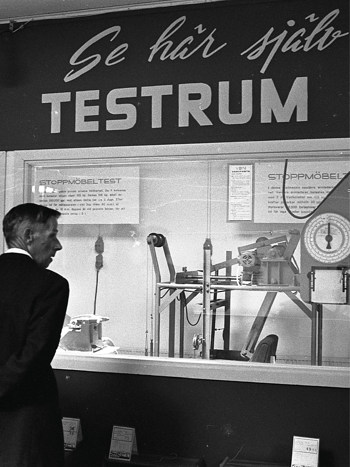 Onder bord 'Kijk zelf maar - testruimte' kijken twee mannen door glas naar machines die testen hoe lang de meubels meegaan.