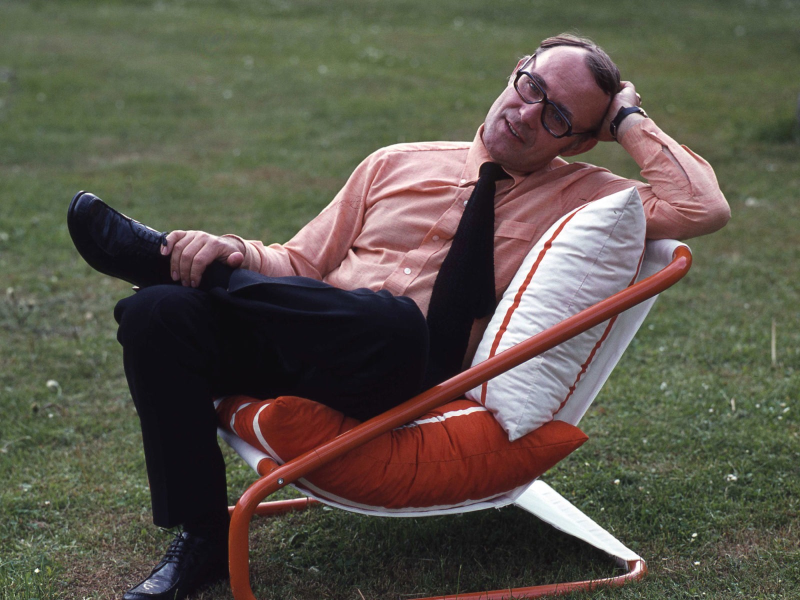 Ingvar Kamprad i rosa skjorta och svart slips, avkopplande i en röd och vit stol på 1970-talet på en gräsmatta.