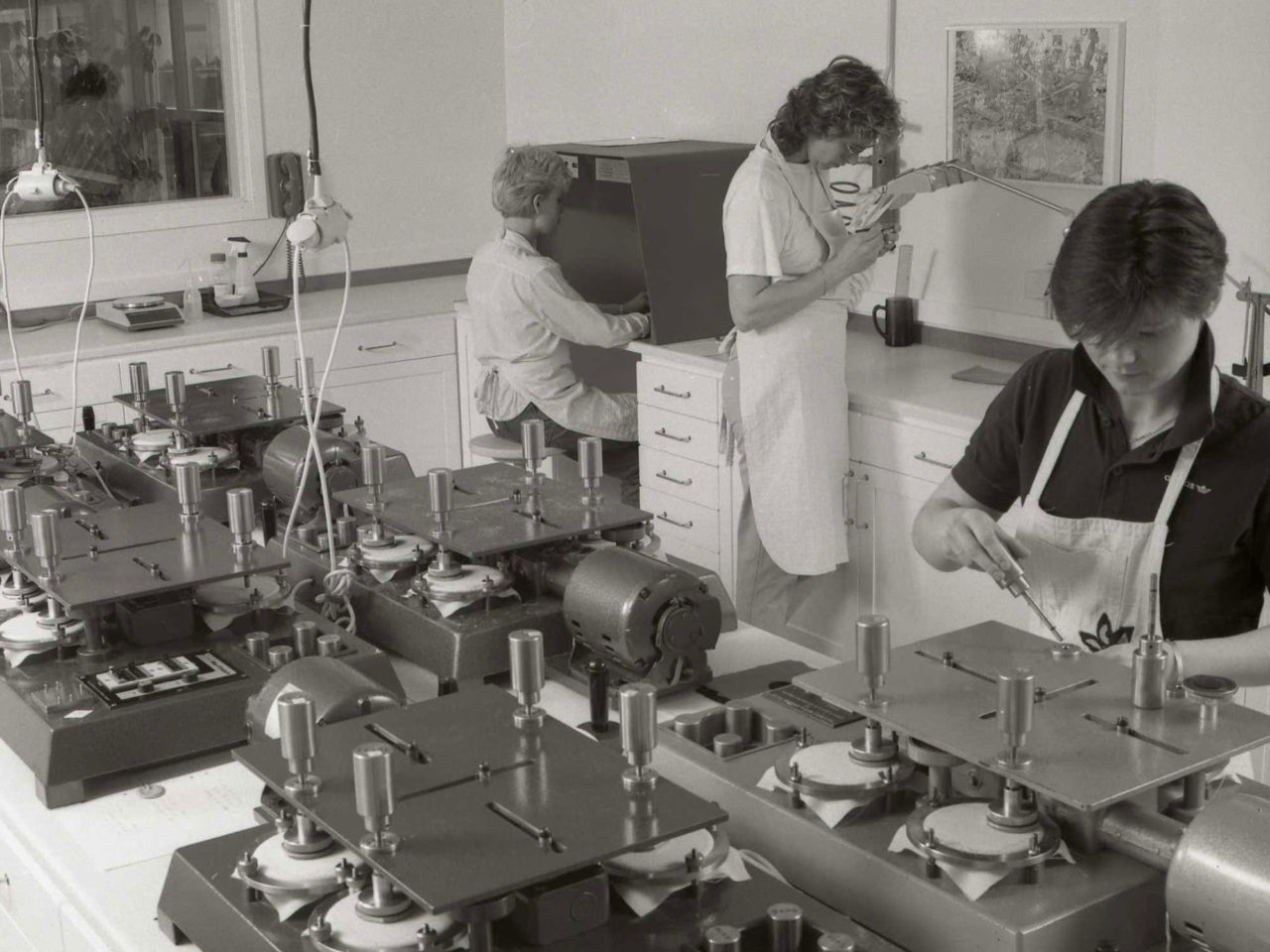 Drie vrouwen die dingen controleren in een testlab, met machines of onder sterke lampen en vergrootglazen.