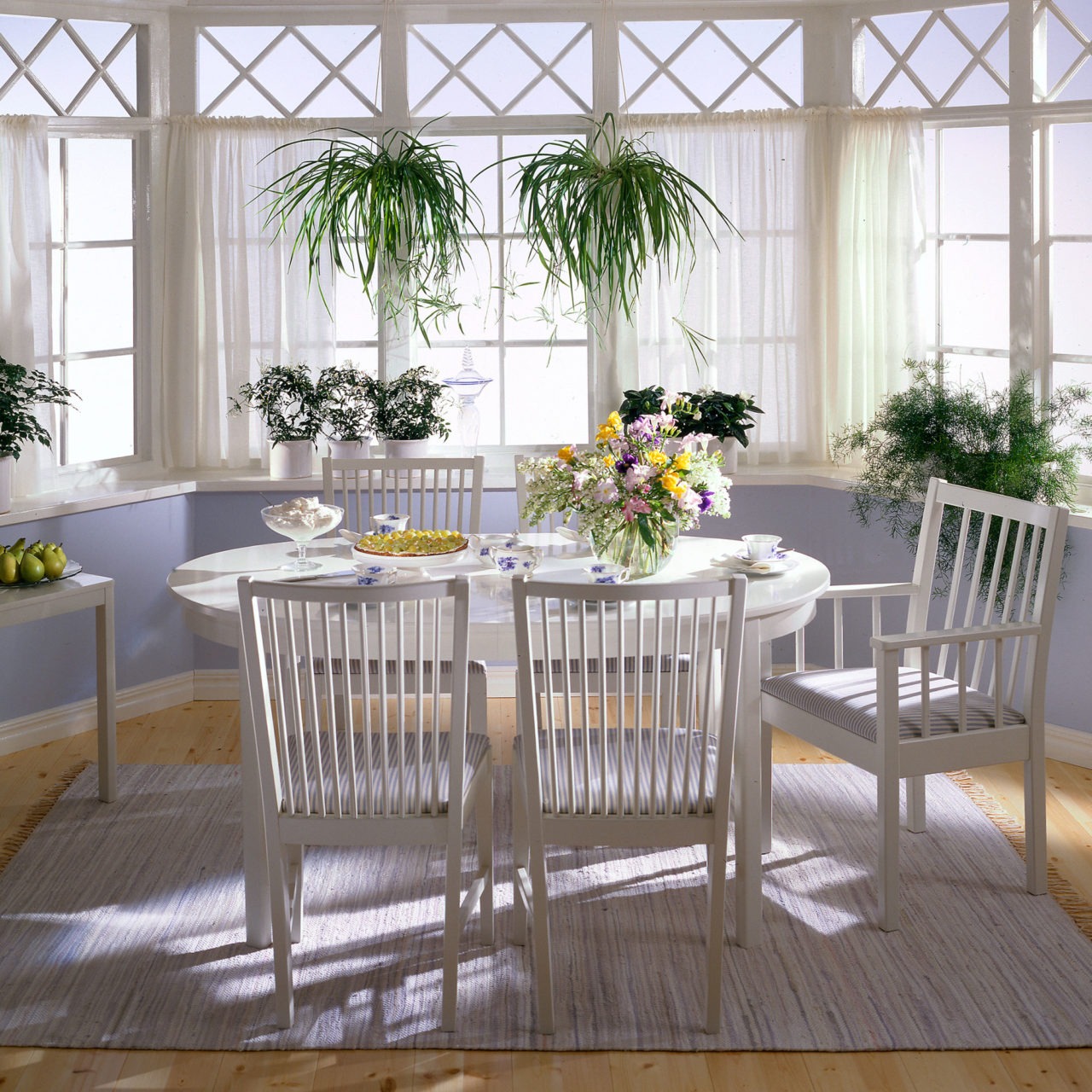 Pin-back chairs with and without armrests around an elliptical dining table, all white in model STOCKHOLM, in a light room with a bay windows.
