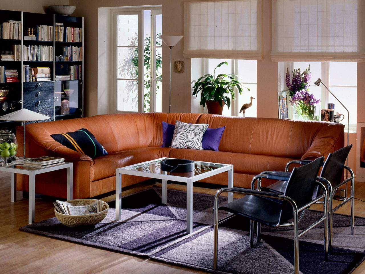 A living room with parquet floors and a long corner sofa in brown leather. Nearby are two chairs in chrome-plated metal and black leather.