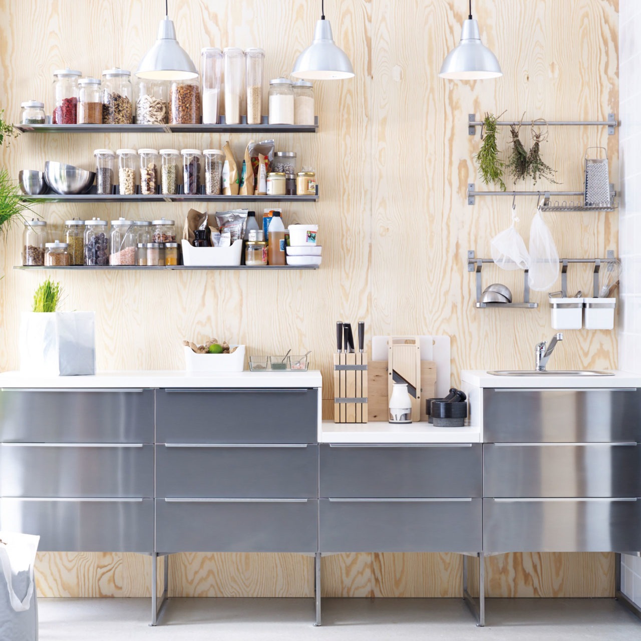 Cocina modular con cajoneras en metal cepillado y pared de madera contrachapada. Las estanterías contienen alimentos secos.