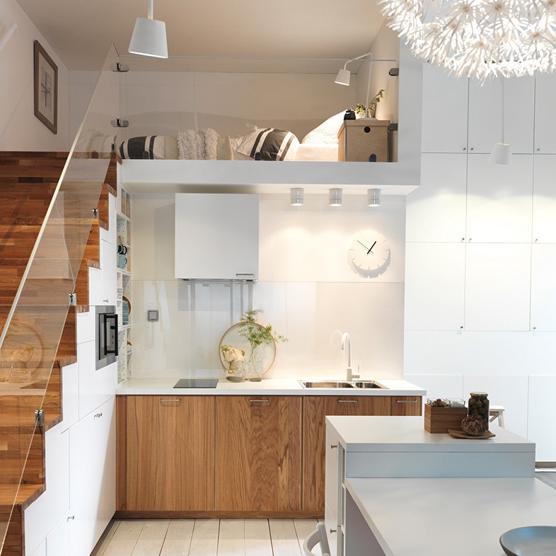Cocina junto a escalera que lleva a un loft para dormir. Armarios y escalones revestidos de madera; el resto es blanco.