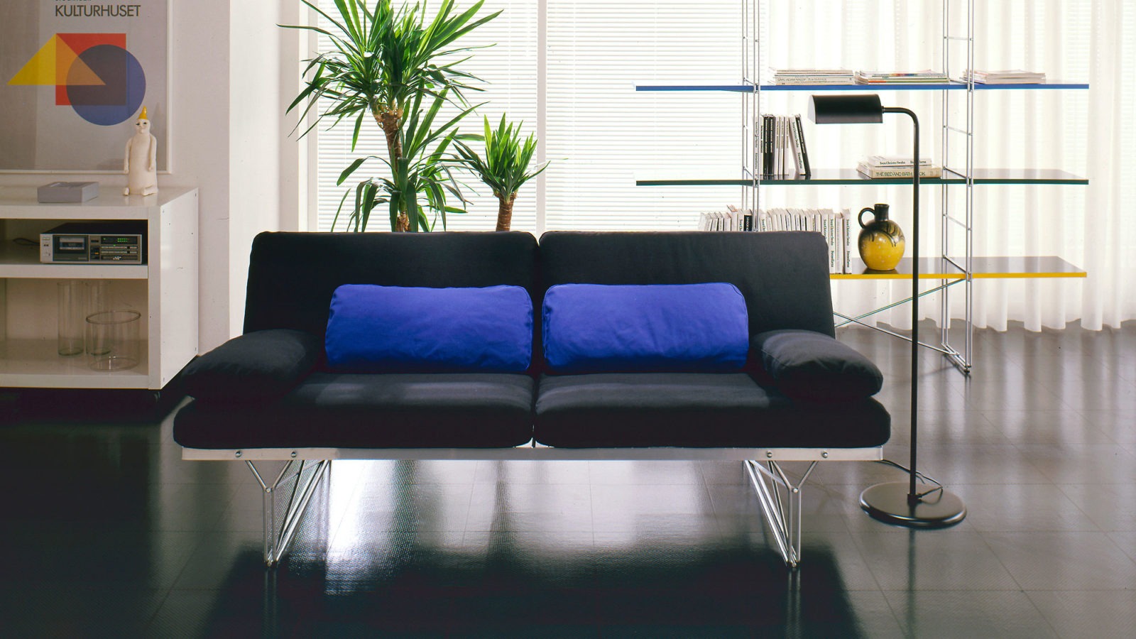 Sofá negro MOMENT y estantería multicolor MOMENT con fina estructura metálica en una habitación de brillante suelo negro.